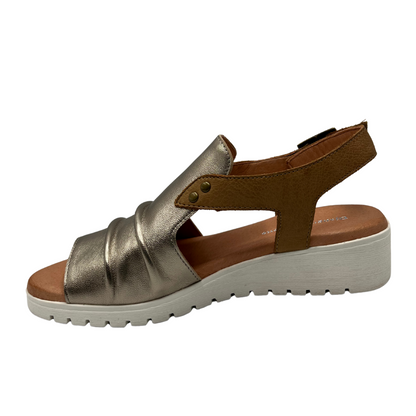 Left facing view of leather sandal with a champagne gold upper and adjustable tan leather strap. Low wedge heel and rounded peep toe.