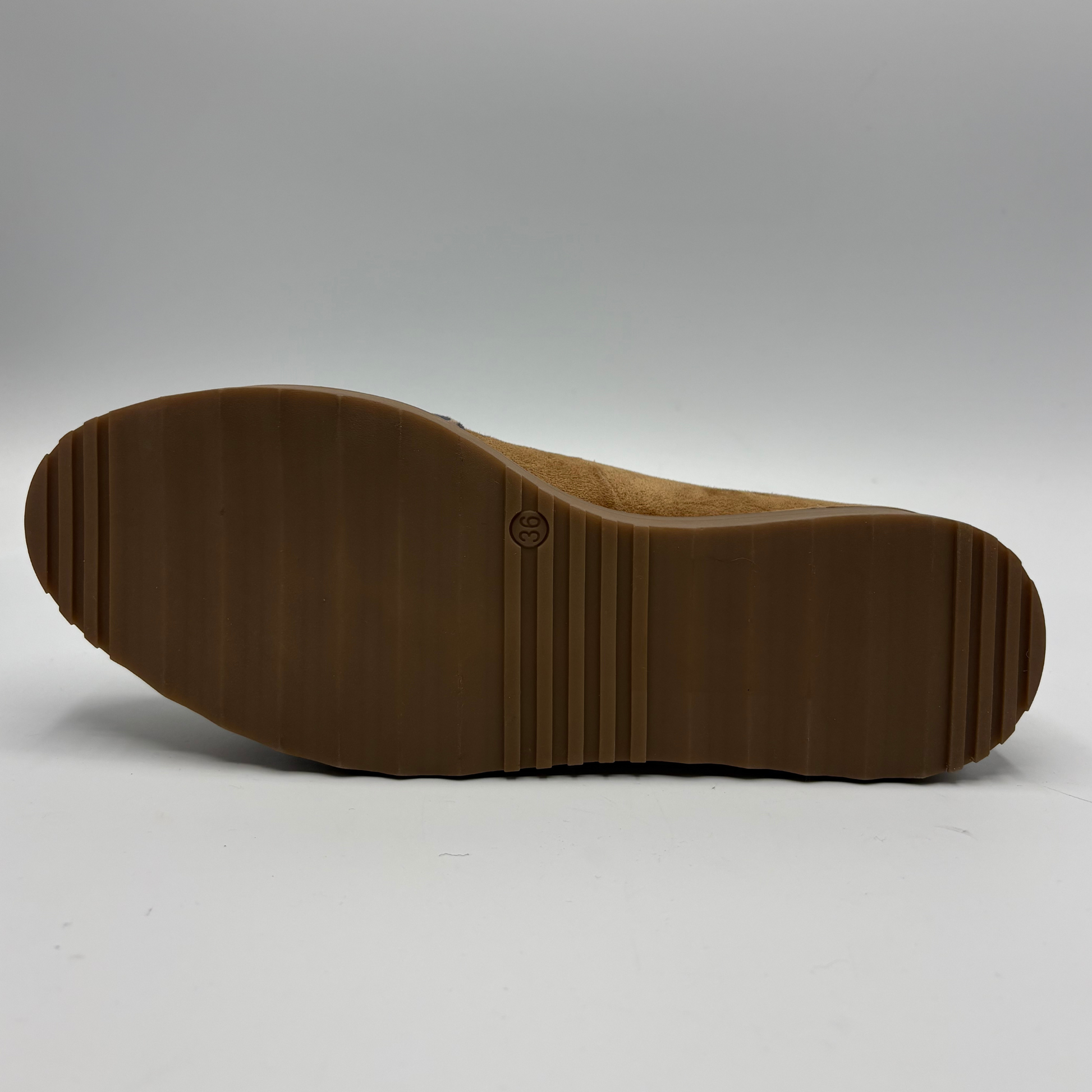 The outsole of a brown suede shoe, showing a textured lug pattern and a small embossed circular logo, photographed against a neutral gray background.