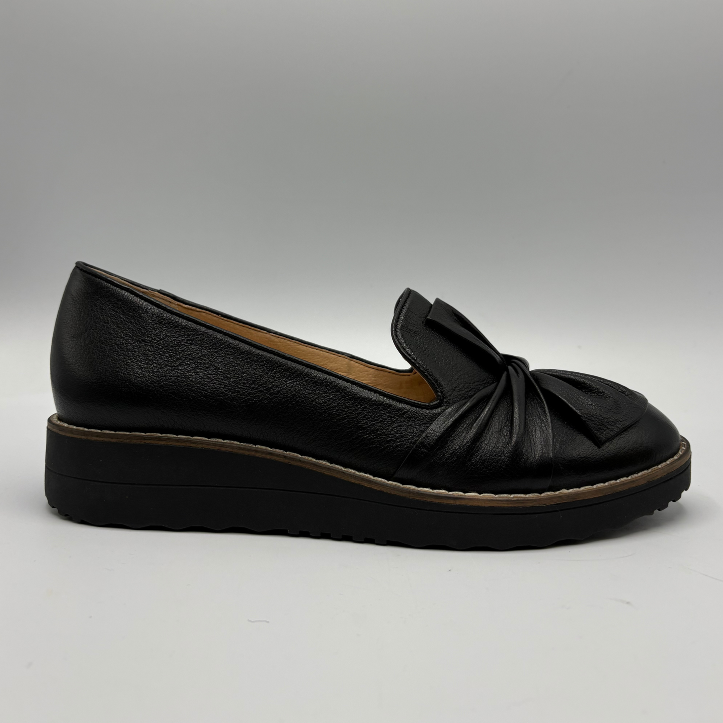 A single black leather slip-on loafer with a decorative bow detail on the vamp and a thick black platform sole, shown against a neutral gray background.