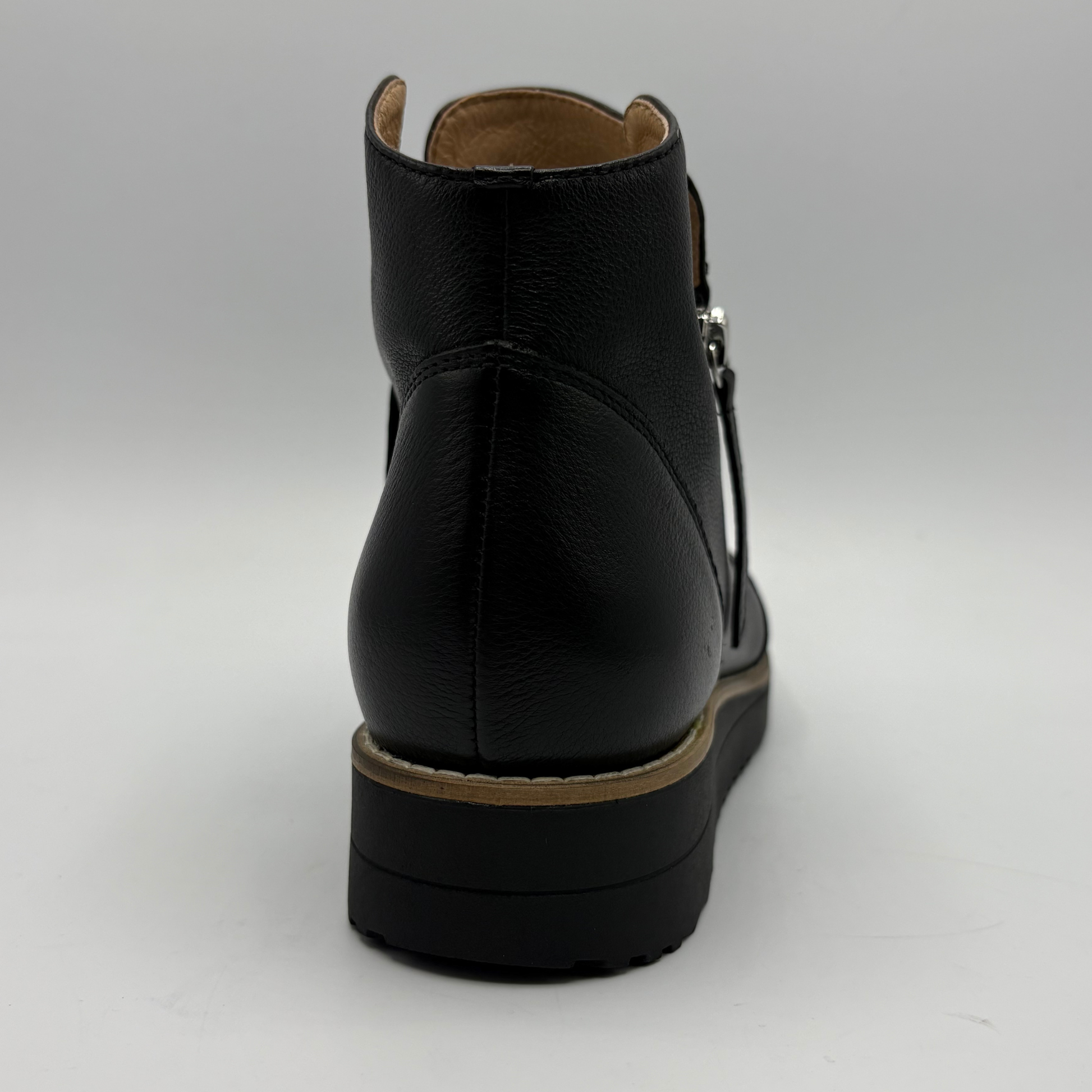 A black leather ankle boot shown from the back, featuring a low black platform sole with a tan stripe, a tan interior lining, and a silver zipper pull on a light gray background.