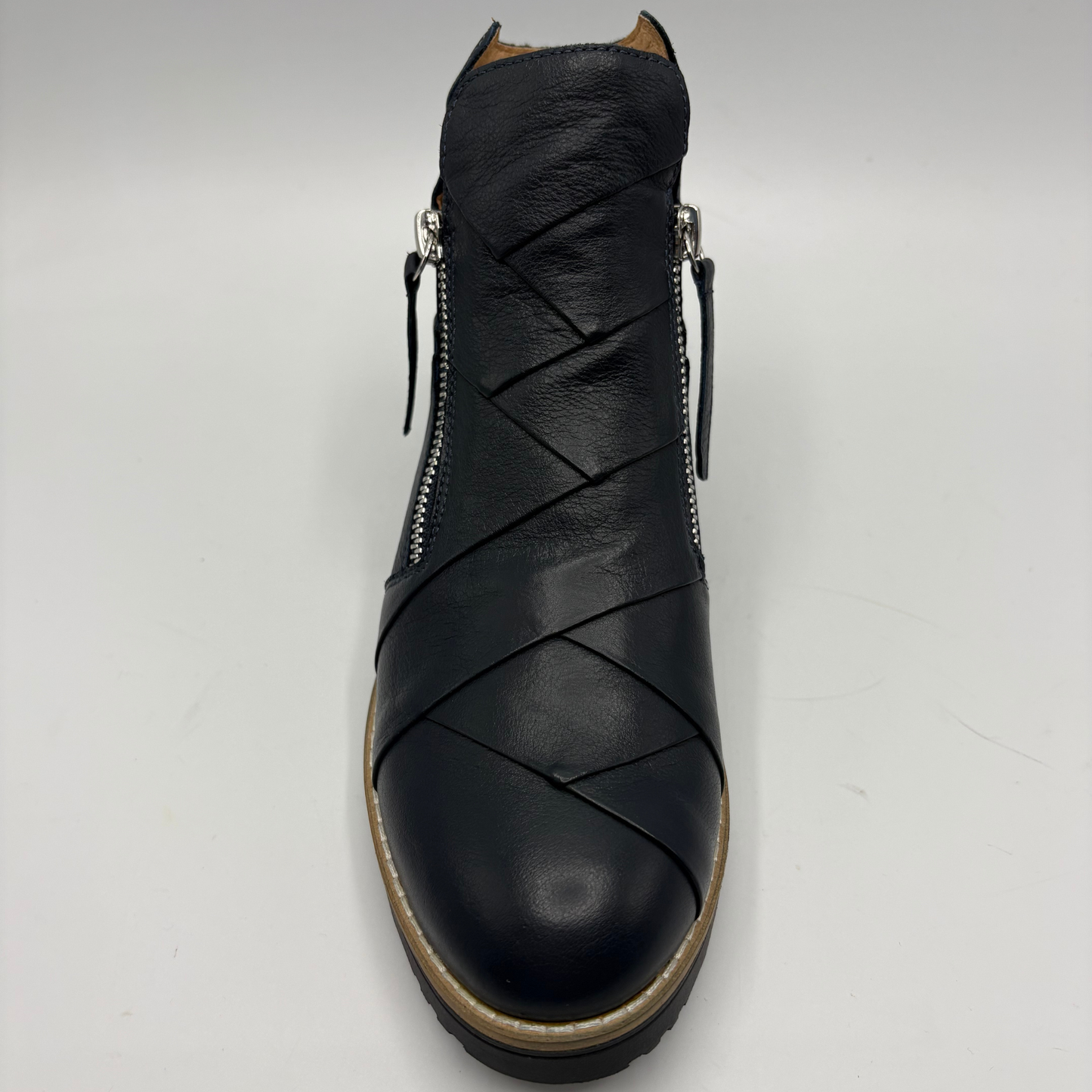 A black leather ankle boot with diagonal wrap-around straps across the vamp, side zippers, and a tan outsole, shown from a front-facing perspective on a light gray background.