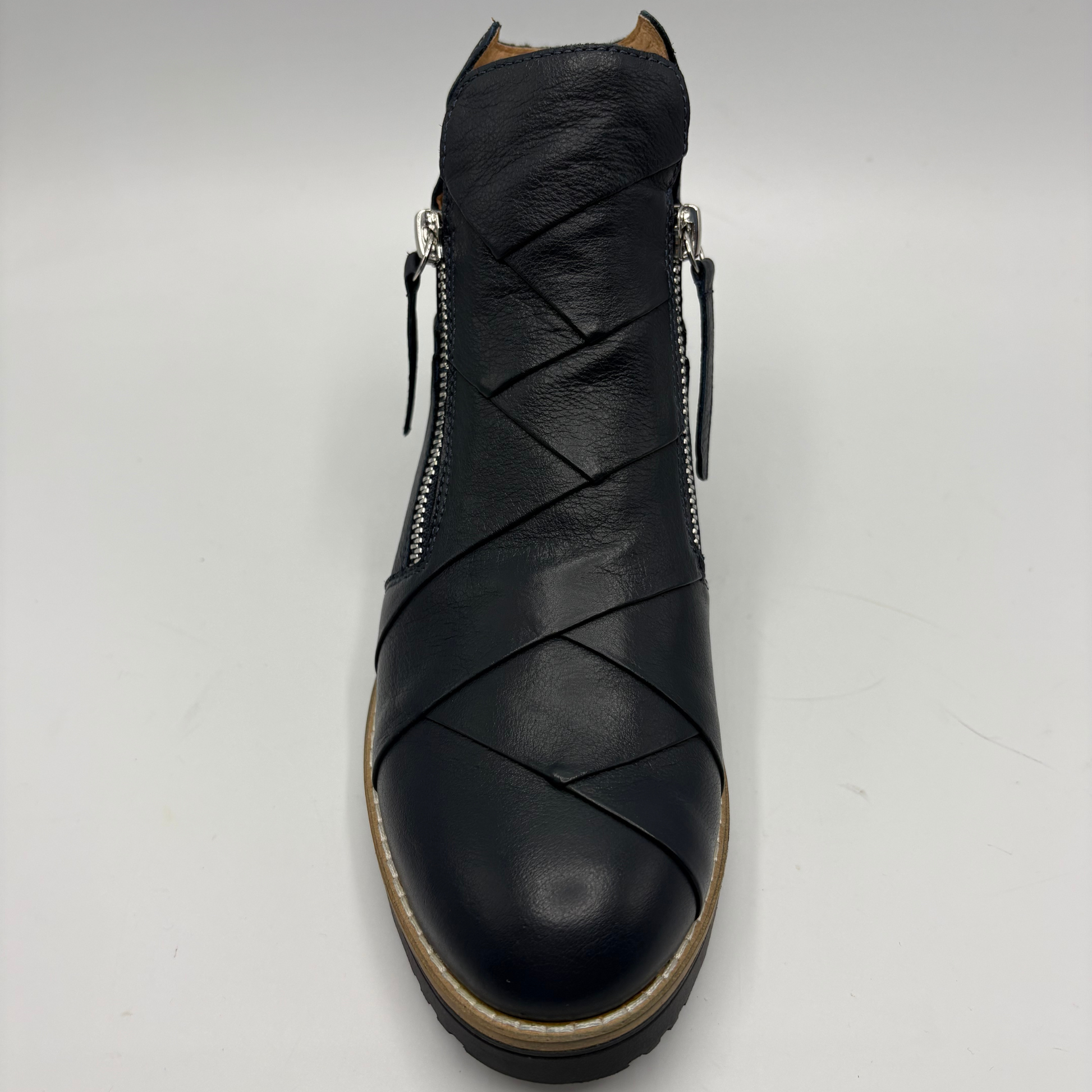 A black leather ankle boot with diagonal wrap-around straps across the vamp, side zippers, and a tan outsole, shown from a front-facing perspective on a light gray background.