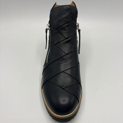 A black leather ankle boot with diagonal wrap-around straps across the vamp, side zippers, and a tan outsole, shown from a front-facing perspective on a light gray background.