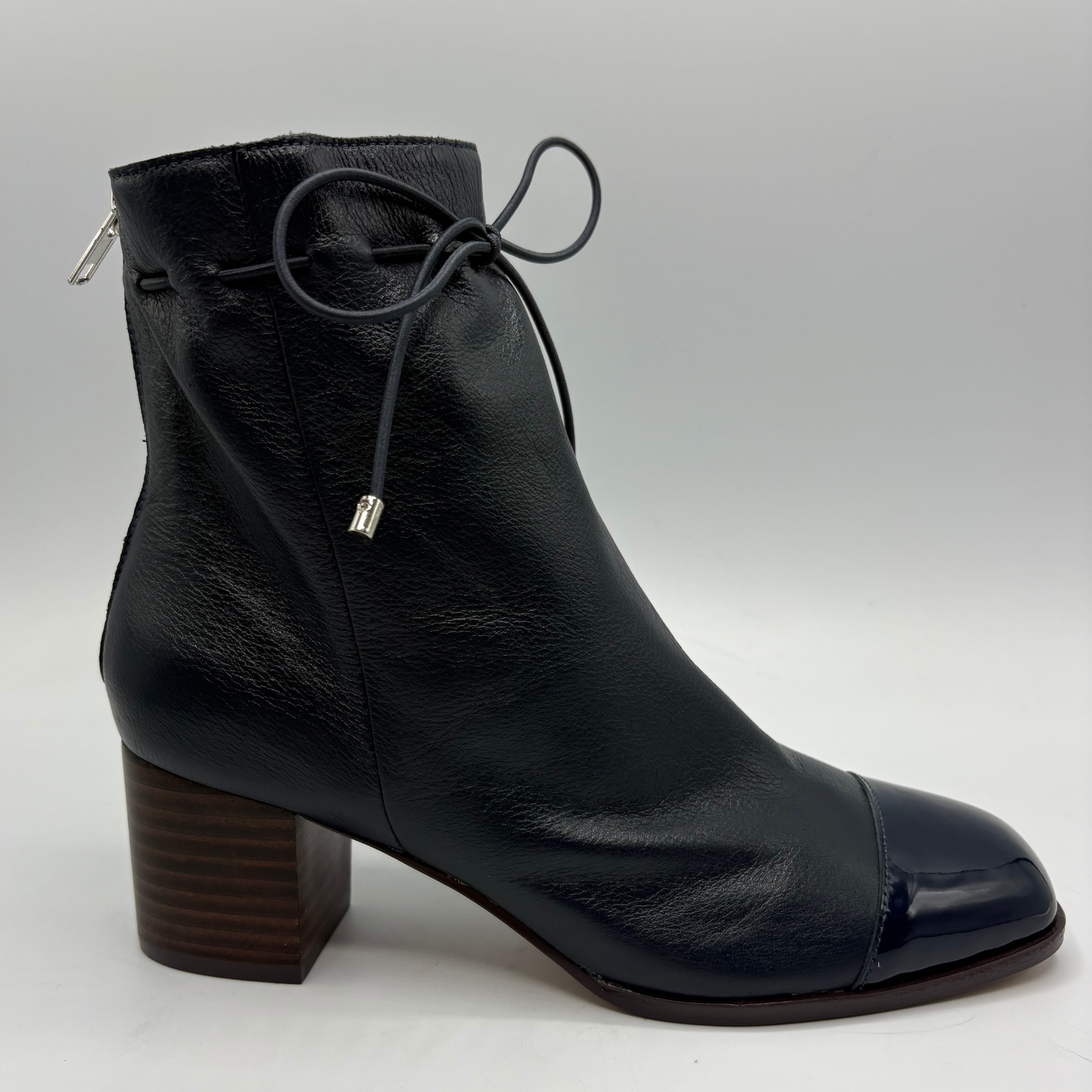 A navy leather ankle boot with a chunky brown block heel, a glossy cap toe, and thin black laces tied at the ankle, shown against a light gray background.