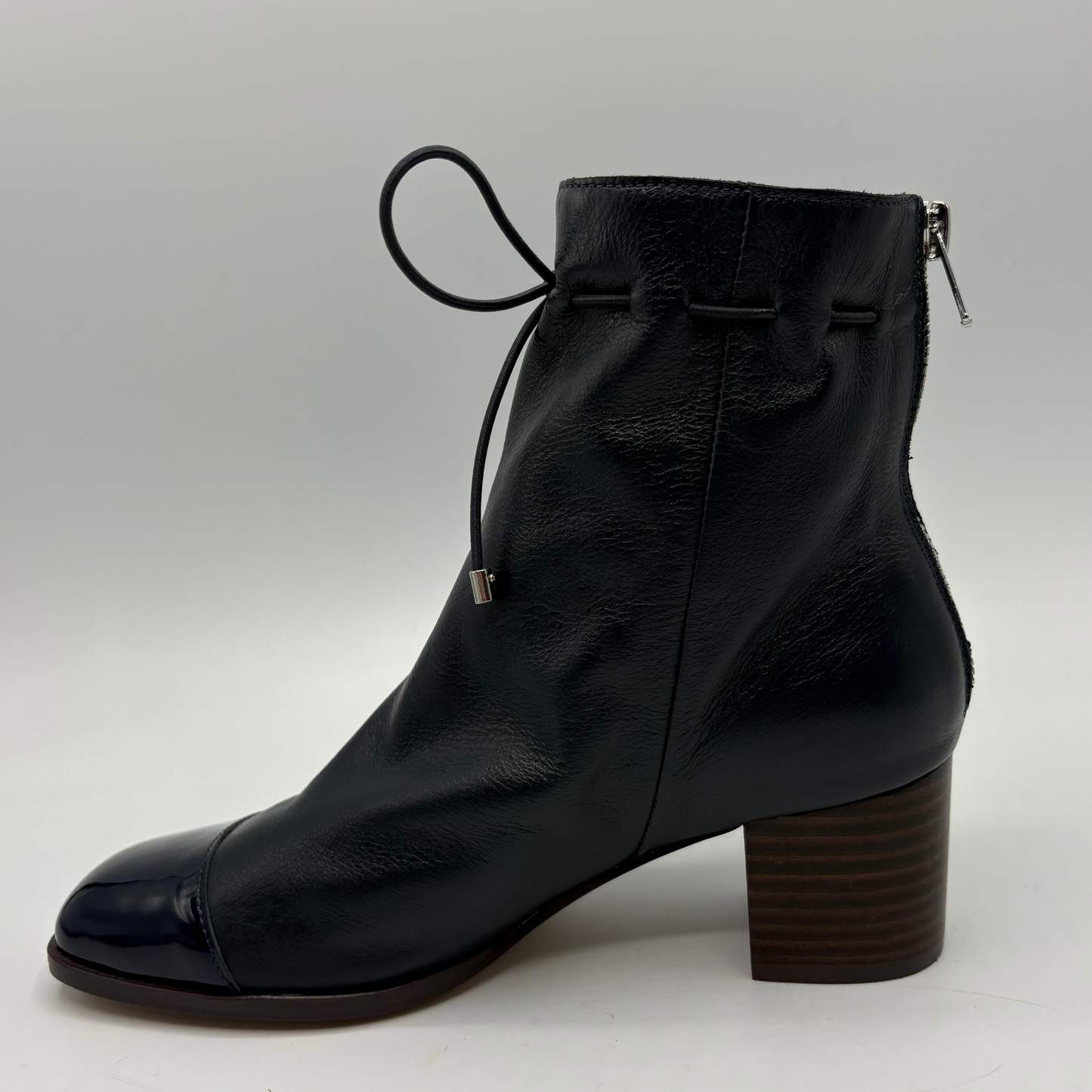 A navy leather ankle boot with a side zipper, a decorative hanging tie detail, and a stacked block heel, shown against a neutral light gray background.