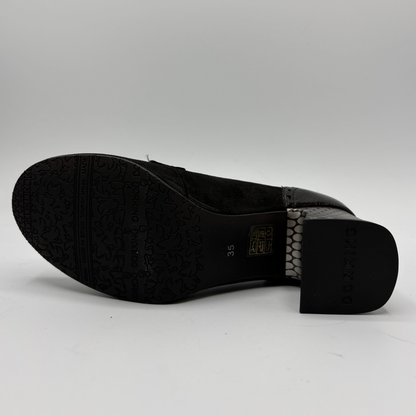 A black high-heeled shoe shown from a side angle, displaying its textured black sole, a gold brand plaque, and a black and white patterned heel counter on a light gray background.