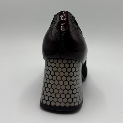 A close-up rear view of a black high-heeled shoe with a glossy patent finish and a chunky block heel covered in a silver-gray circular honeycomb pattern, set against a plain light gray background.