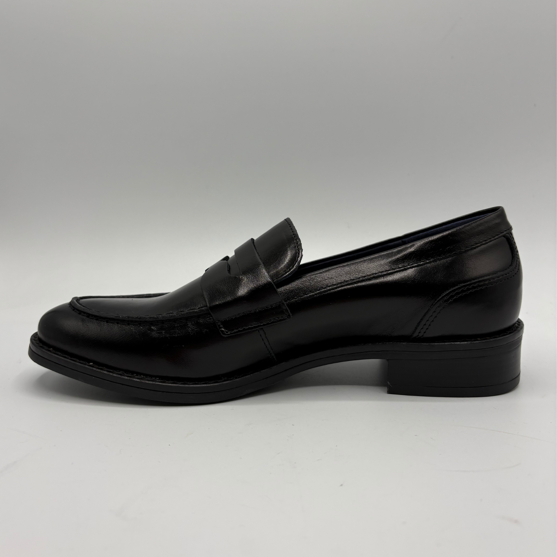 A single black leather penny loafer with a low block heel, shown in a side profile view against a neutral gray background.