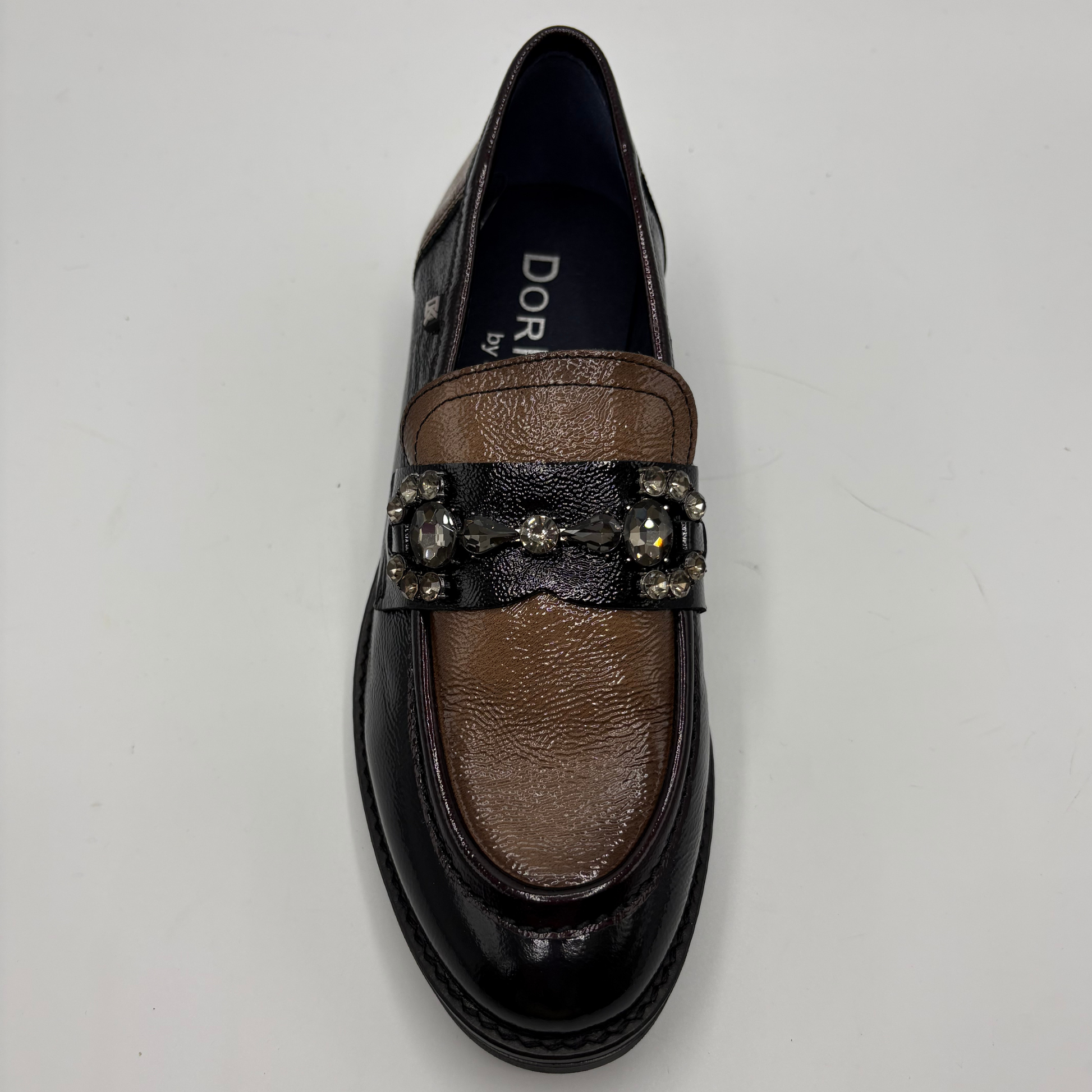 A single black leather loafer with a brown textured leather panel across the vamp, embellished with a black strap and decorative crystal-like hardware, photographed from above on a light gray background.