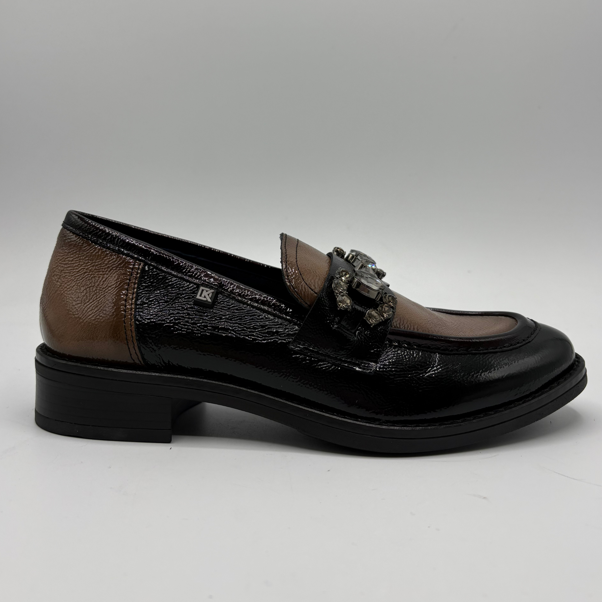 A women's loafer-style shoe in black patent leather and brown textured leather, featuring a decorative metallic buckle detail on the strap, set against a neutral gray background.