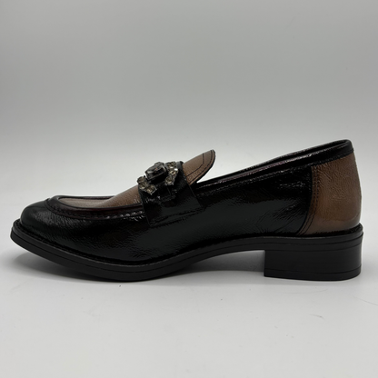 A single women's loafer-style shoe shown in side profile on a light gray studio background. The shoe has a glossy black toe and heel, a medium brown leather side panel, and a small metallic decorative embellishment across the vamp.