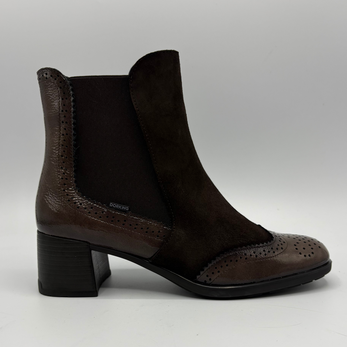 A dark brown ankle boot with a mix of textured leather and suede panels, a black elastic side panel, and a chunky black block heel, shown from a side profile on a light gray background.