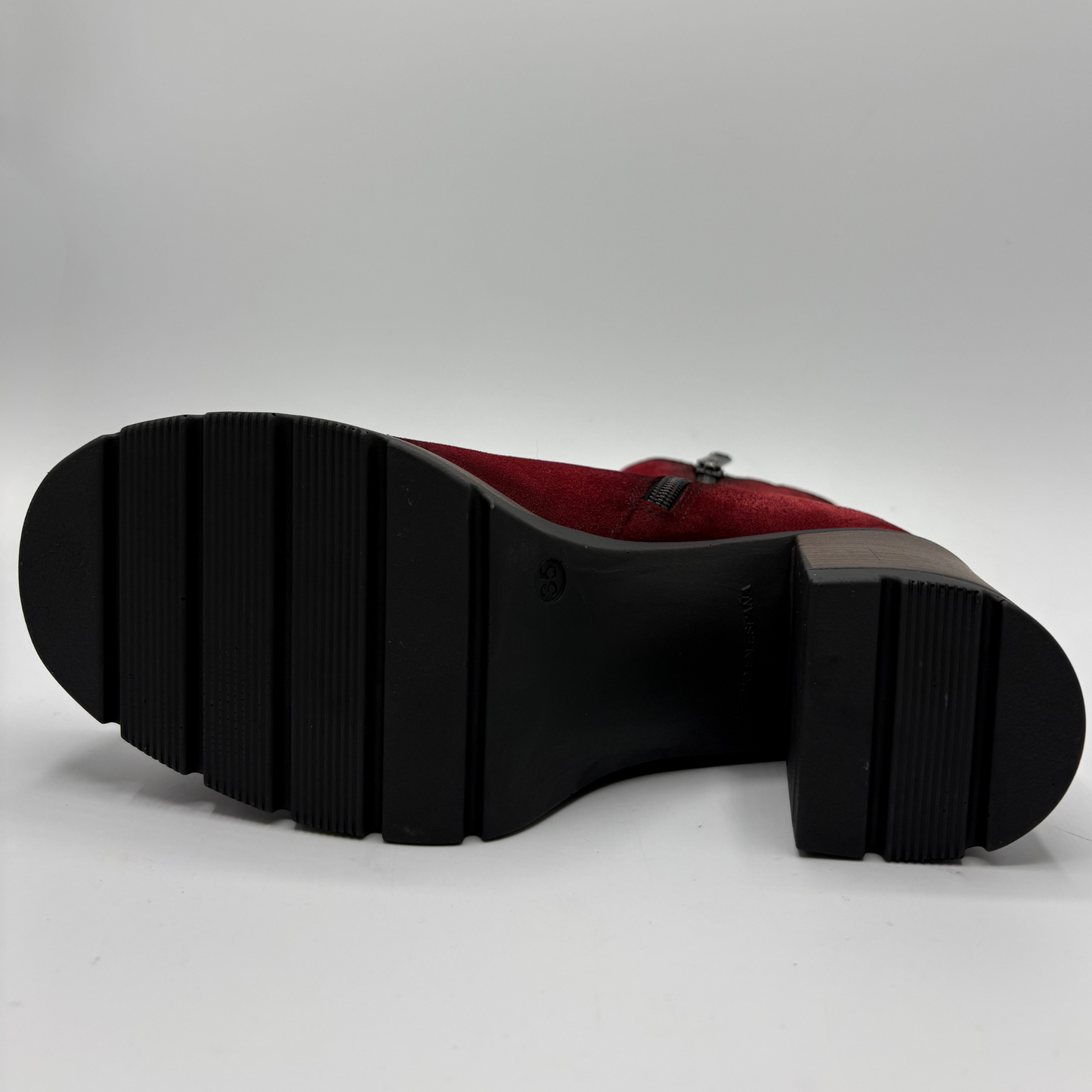 A close-up shot of the black treaded sole of a red suede boot against a light gray background.