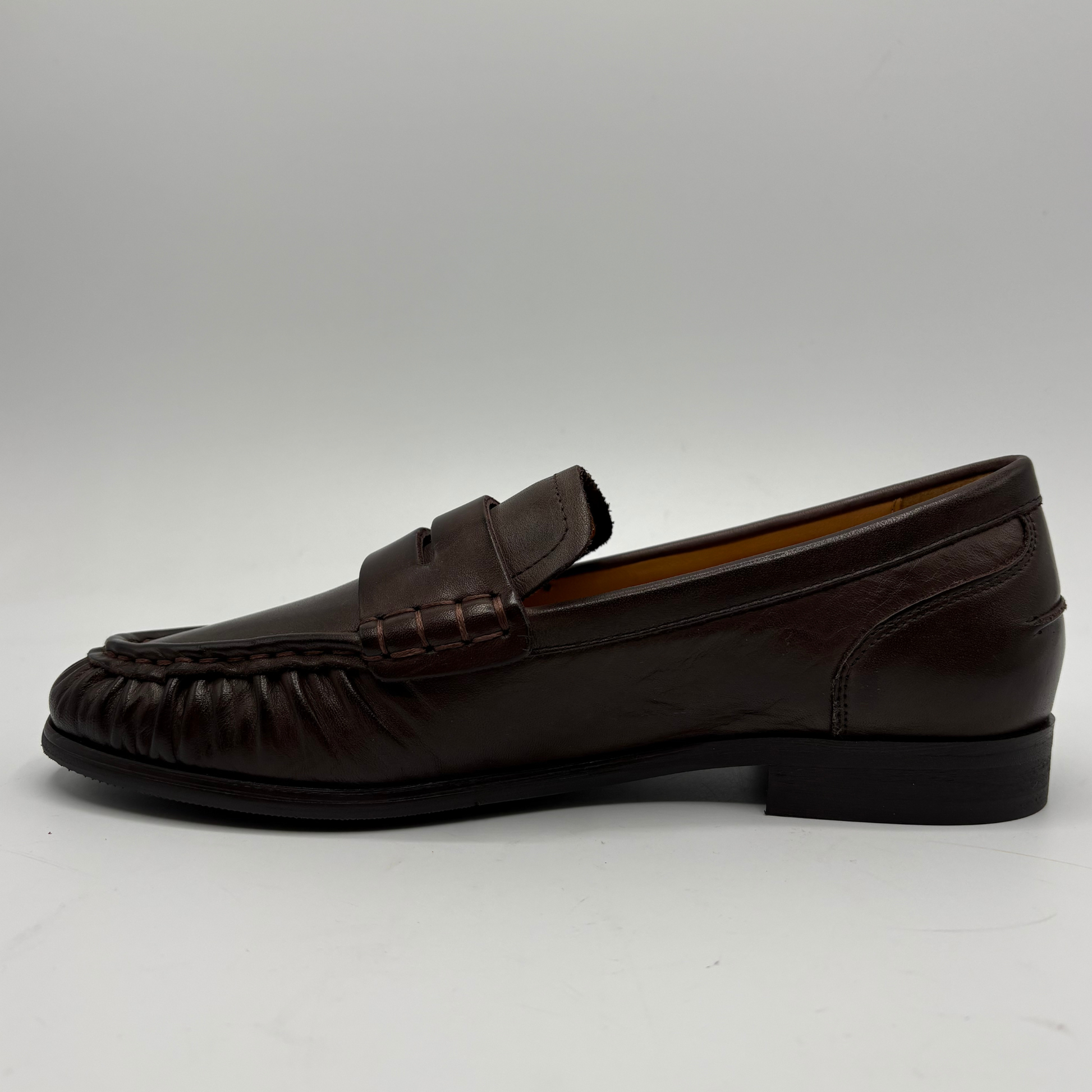 A dark brown leather penny loafer shown from a side profile view against a neutral gray background.