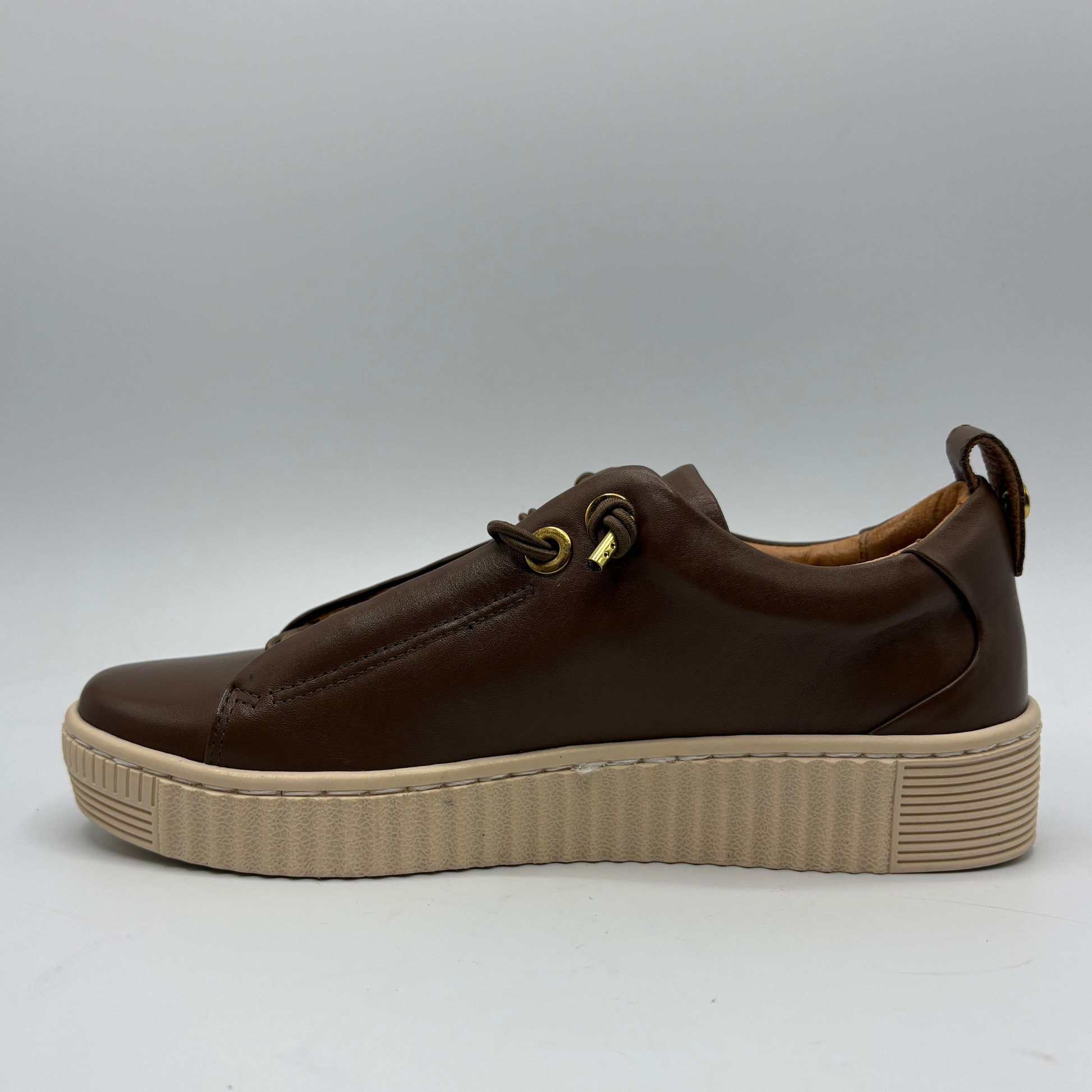A dark brown leather low-top sneaker with a thick beige platform sole, featuring decorative gold-tone eyelets and a pull tab on the heel, shown against a neutral gray background.