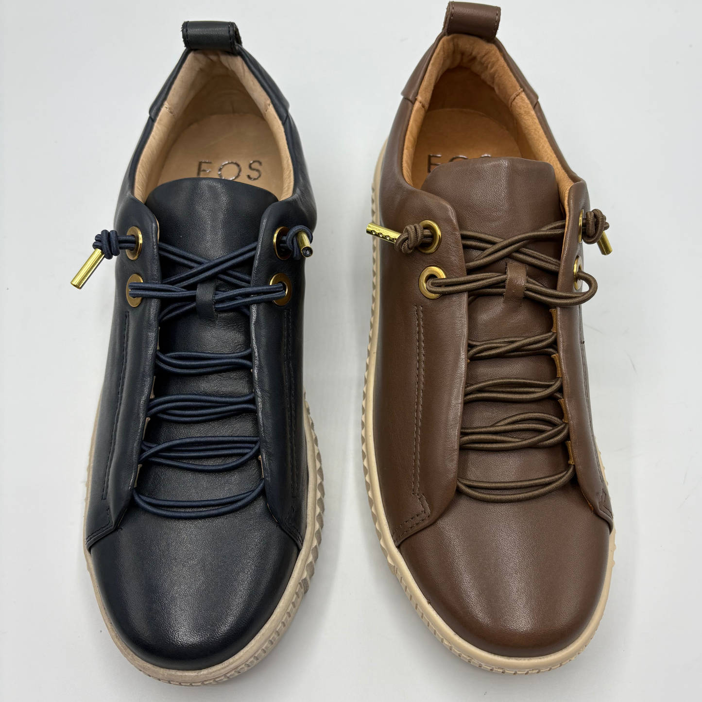 Two low-top leather sneakers shown side by side on a light gray background. The left shoe is dark navy blue with navy laces and gold-tone eyelets, and the right shoe is brown with brown laces and matching gold-tone eyelets.