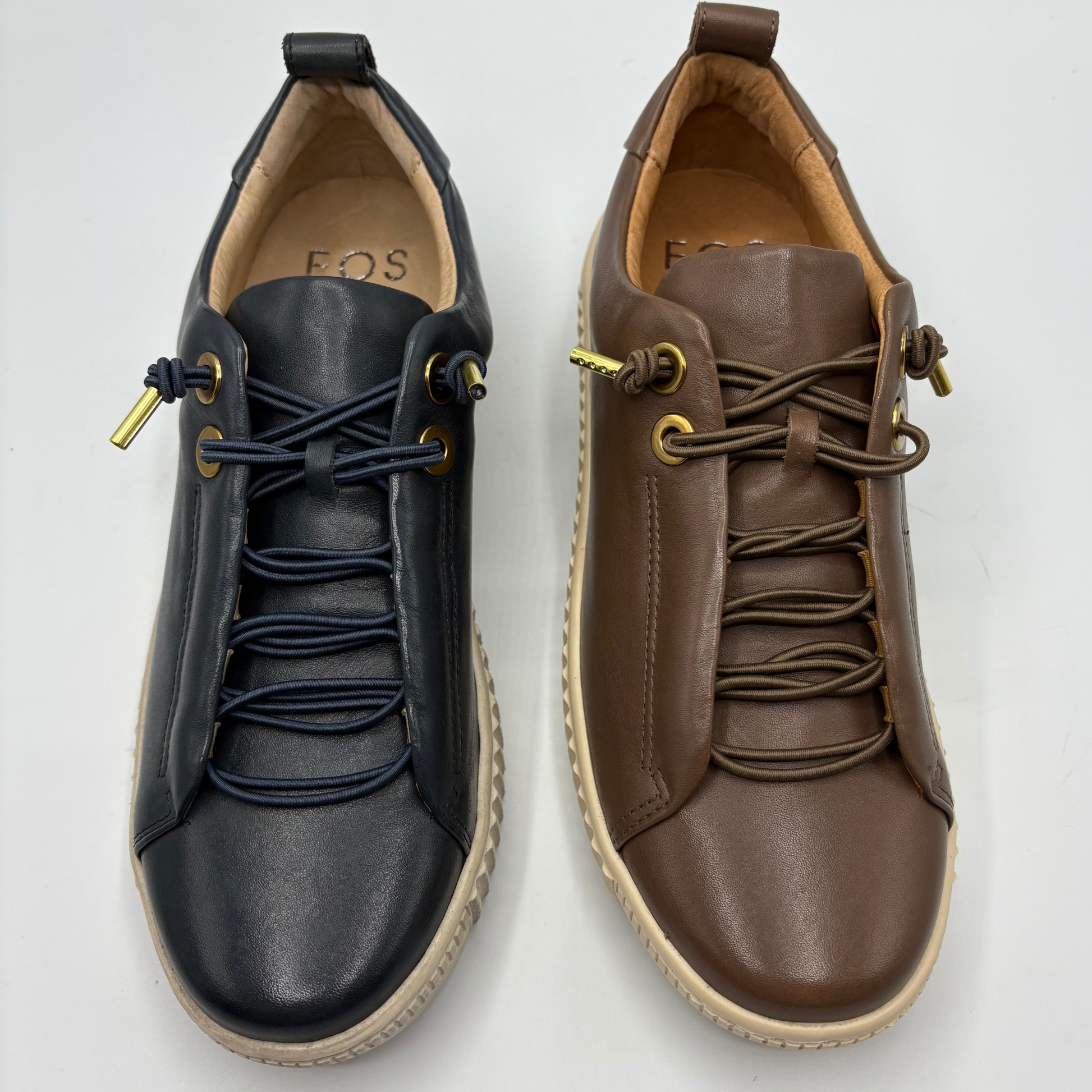 Two low-top leather sneakers shown side by side on a light gray background. The left shoe is dark navy blue with navy laces and gold-tone eyelets, and the right shoe is brown with brown laces and matching gold-tone eyelets.