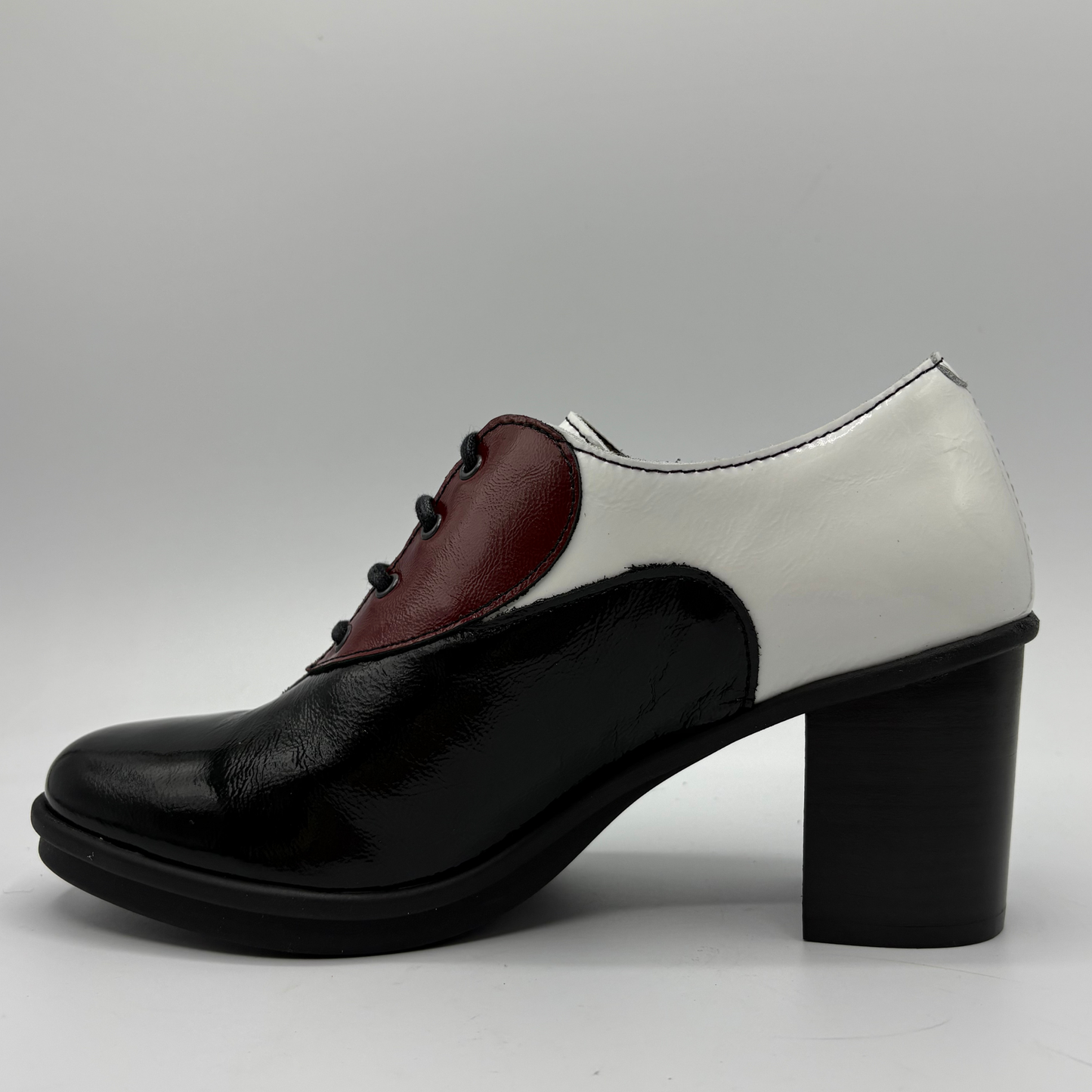 A women's block-heel oxford shoe with a colorblock design, featuring a glossy black toe and heel, a white side panel, and a burgundy leather vamp section with black laces, shown against a neutral gray background.