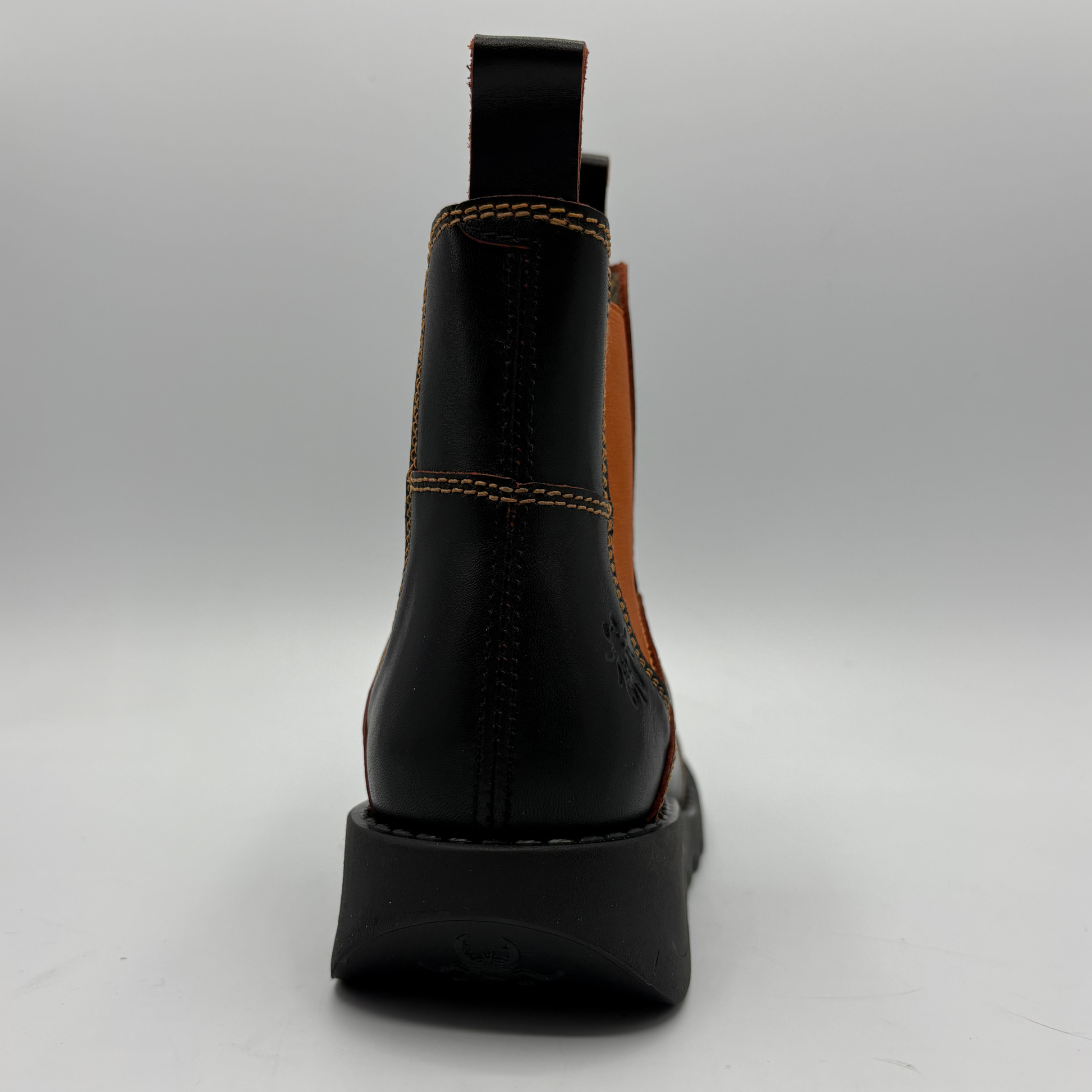 A black leather ankle boot shown from the back, featuring contrast stitching, a tan elastic side panel, and a chunky black sole on a light gray background.