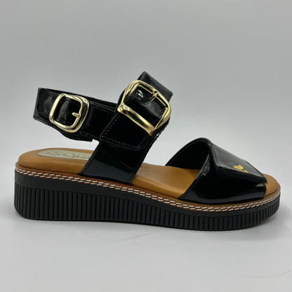 Right facing view of a black patent leather sandal with decorative gold buckles and a wedge heel.