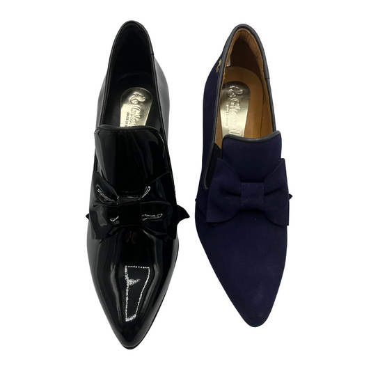 Top view of a pair of heeled loafers side each other. One is black patent leather and the other is dark blue suede. Both have a pointed toe and a bow detail on the upper.