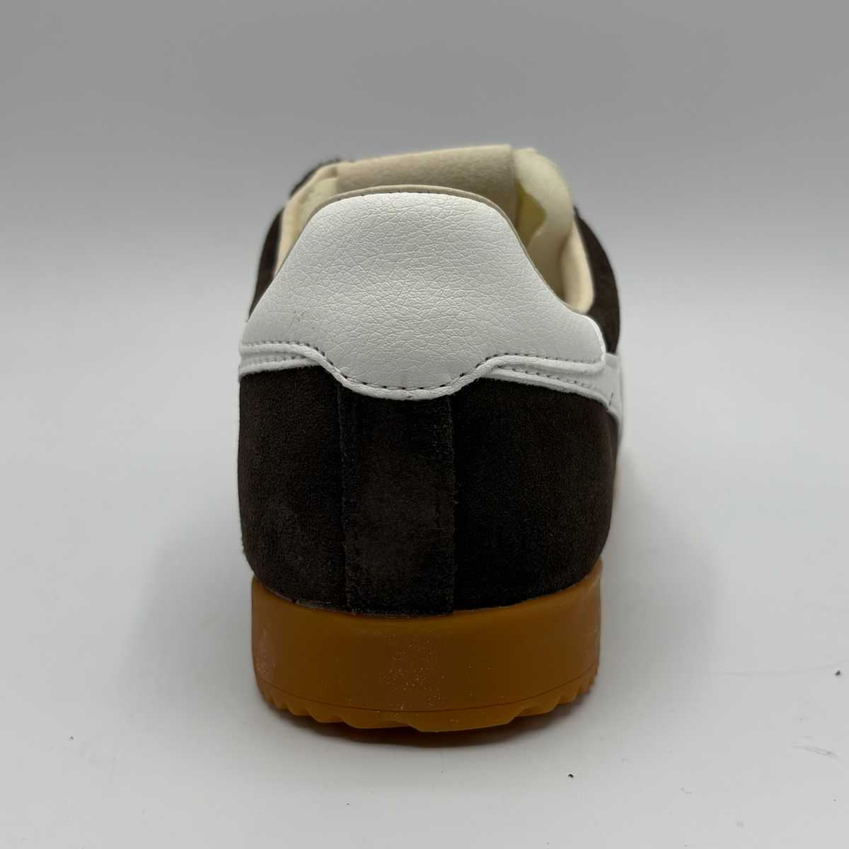 A rear three-quarter view of a low-top sneaker featuring a dark brown suede upper, a white leather heel counter, and a thick brown gum sole, set against a neutral gray background.