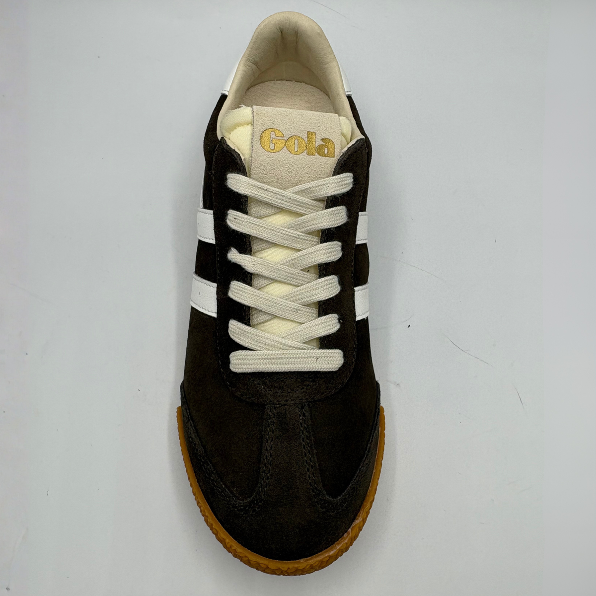 A single dark brown suede sneaker with off-white laces, white side stripes, and a tan rubber sole, photographed from a top-down angle on a light gray background.