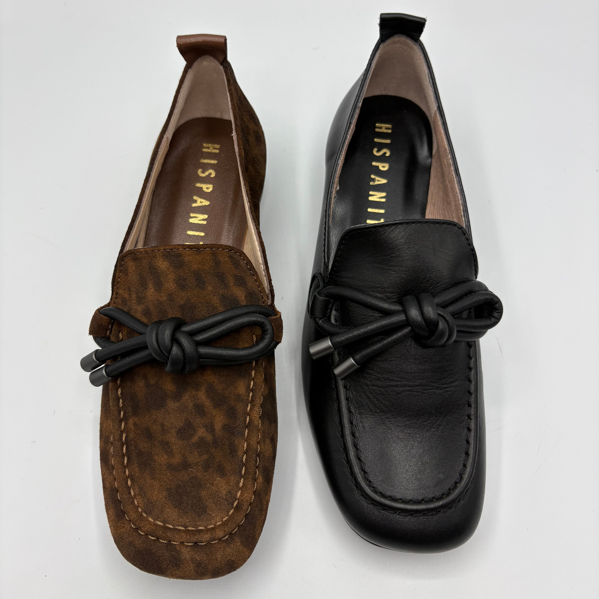 Two women's slip-on loafers displayed side by side on a light gray background. The left loafer is brown with a subtle leopard print pattern and a black knotted strap.