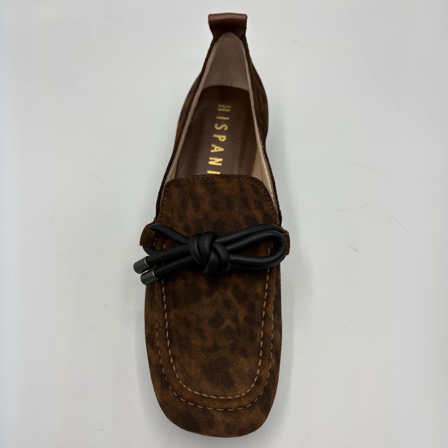 A brown suede loafer with a black leather bow detail across the vamp, photographed from a top-down angle on a light gray background. The brand name 'HISPANITAS' is visible on the insole.
