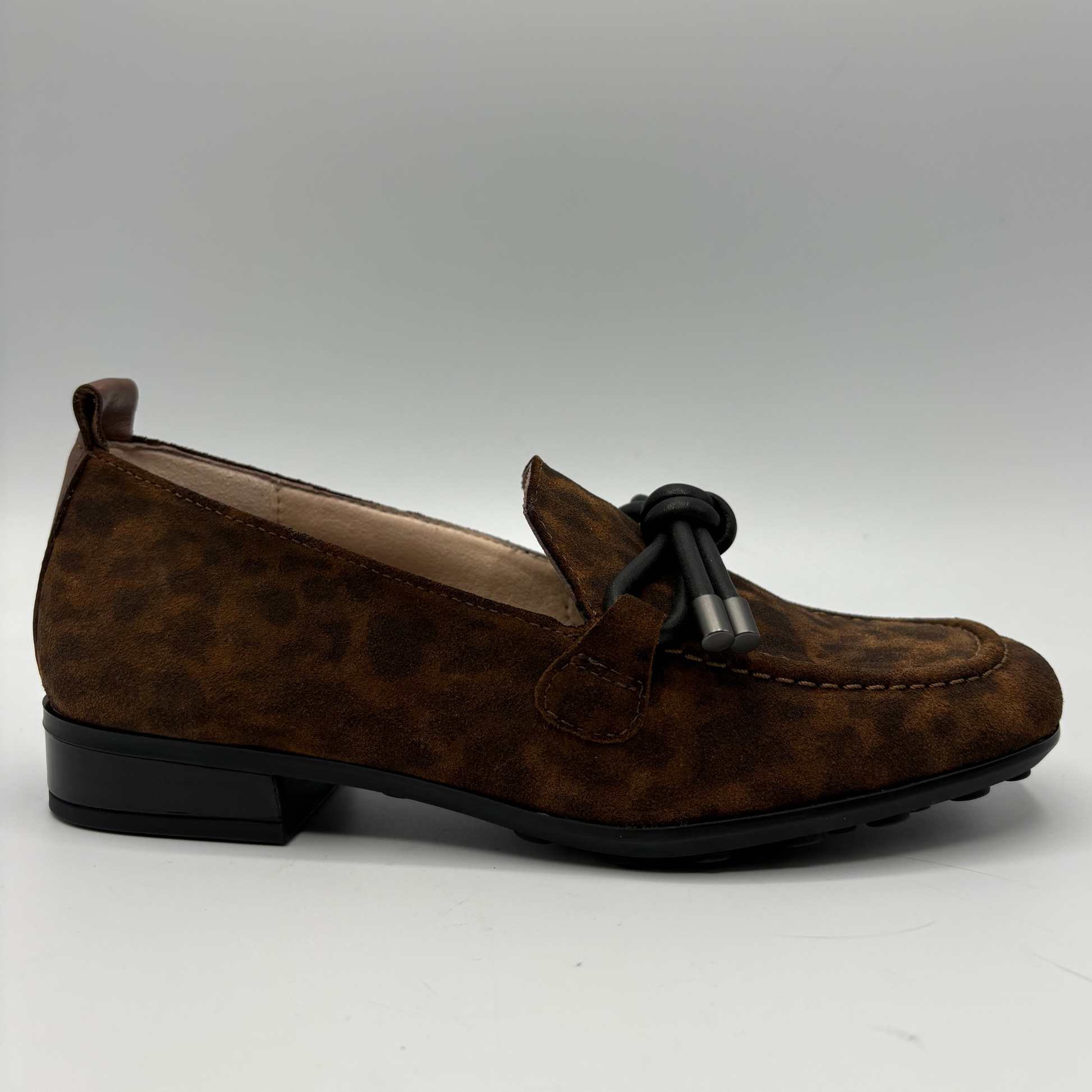 A single brown suede loafer with a black tassel detail on the vamp, photographed from a side profile against a neutral gray background.