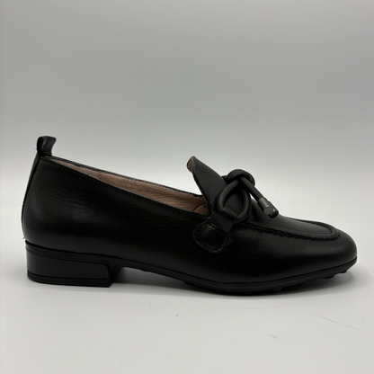 A single black leather loafer with a low block heel and a decorative bow detail on the vamp, photographed against a neutral gray studio background.