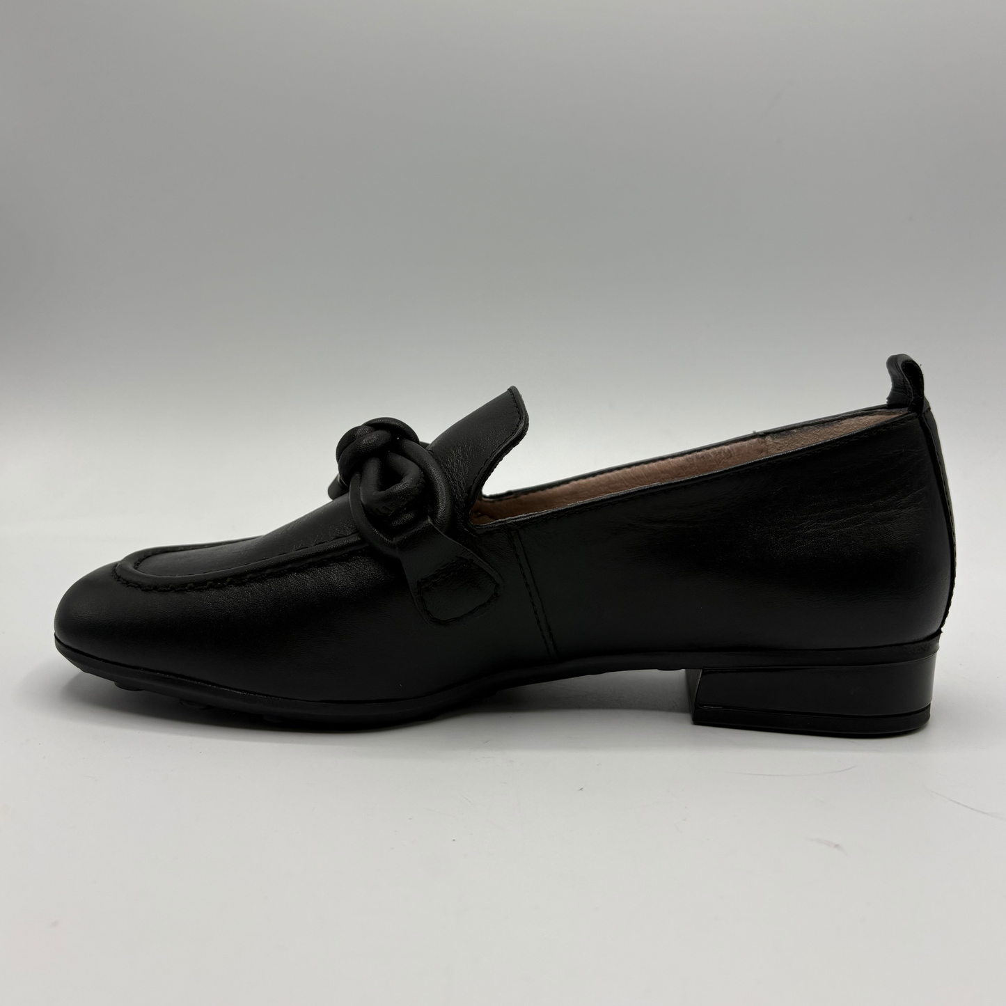 A single black leather loafer with a low block heel and a decorative knotted strap detail across the vamp, shown in profile against a neutral gray background.
