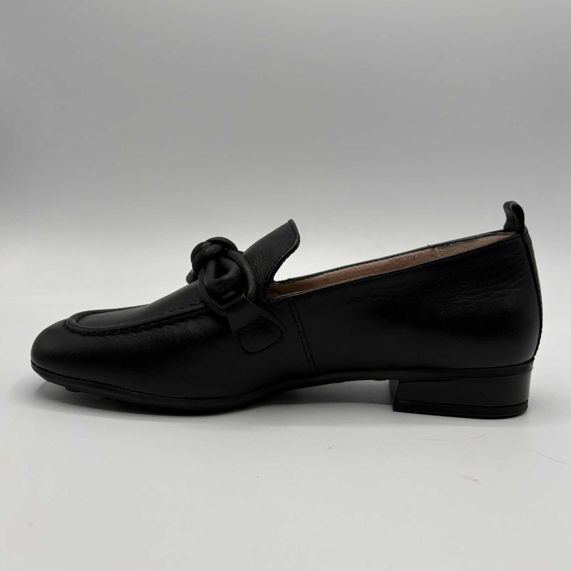 A single black leather loafer with a low block heel and a decorative knotted strap detail across the vamp, shown in profile against a neutral gray background.