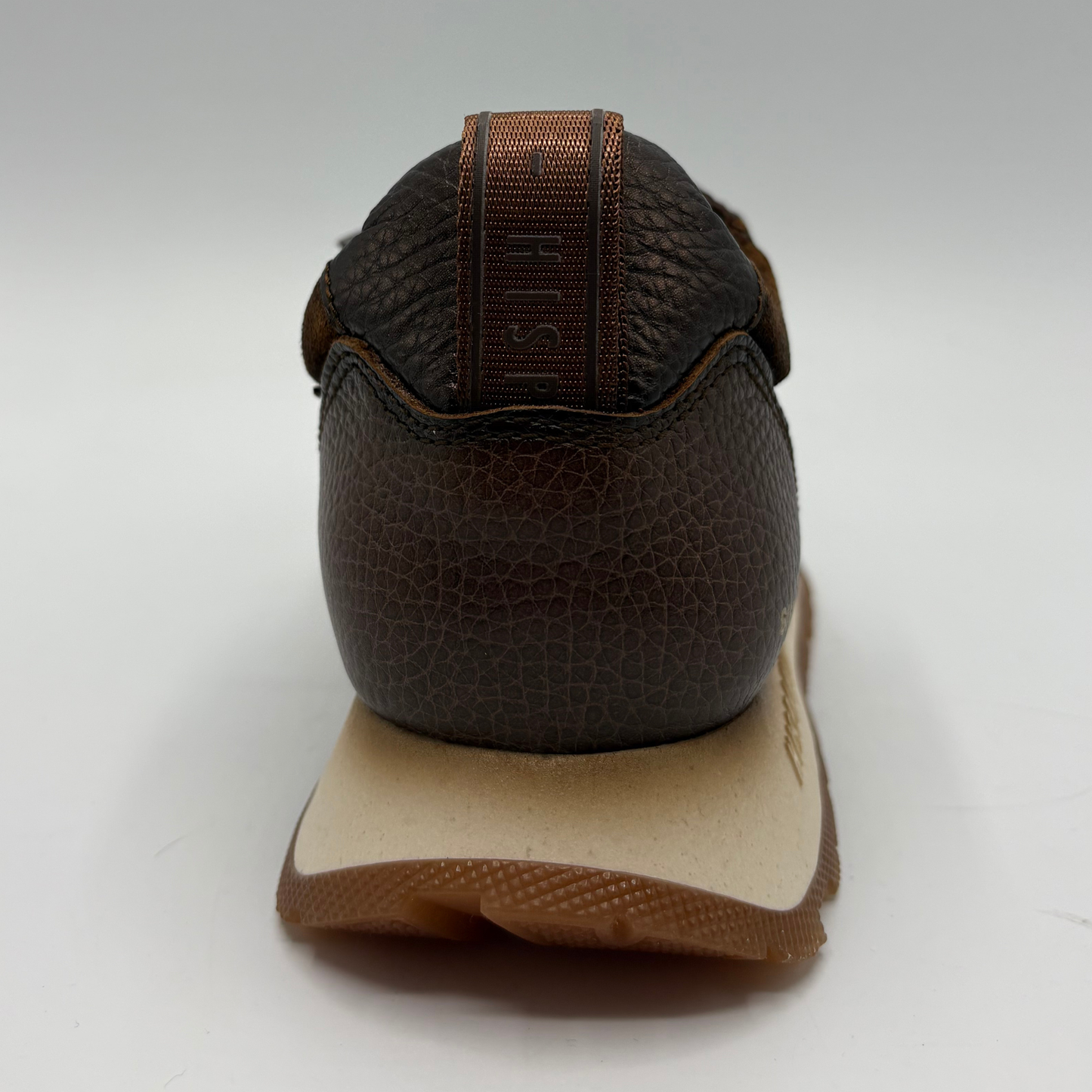 A close-up rear view of a brown textured leather shoe with a thick beige midsole and a gum brown outsole, set against a plain light gray background.