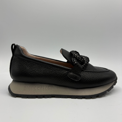 A single black pebbled leather slip-on loafer with a thick braided tassel detail on the vamp and a gray midsole, shown against a neutral gray background. The brand name 'HISPANITAS' is embossed on the side of the gray sole.