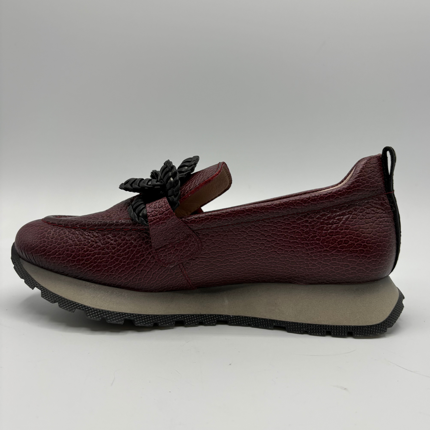 A single burgundy pebbled leather slip-on shoe with a black braided detail on the side and a thick gray and black platform sole, shown against a neutral gray background.
