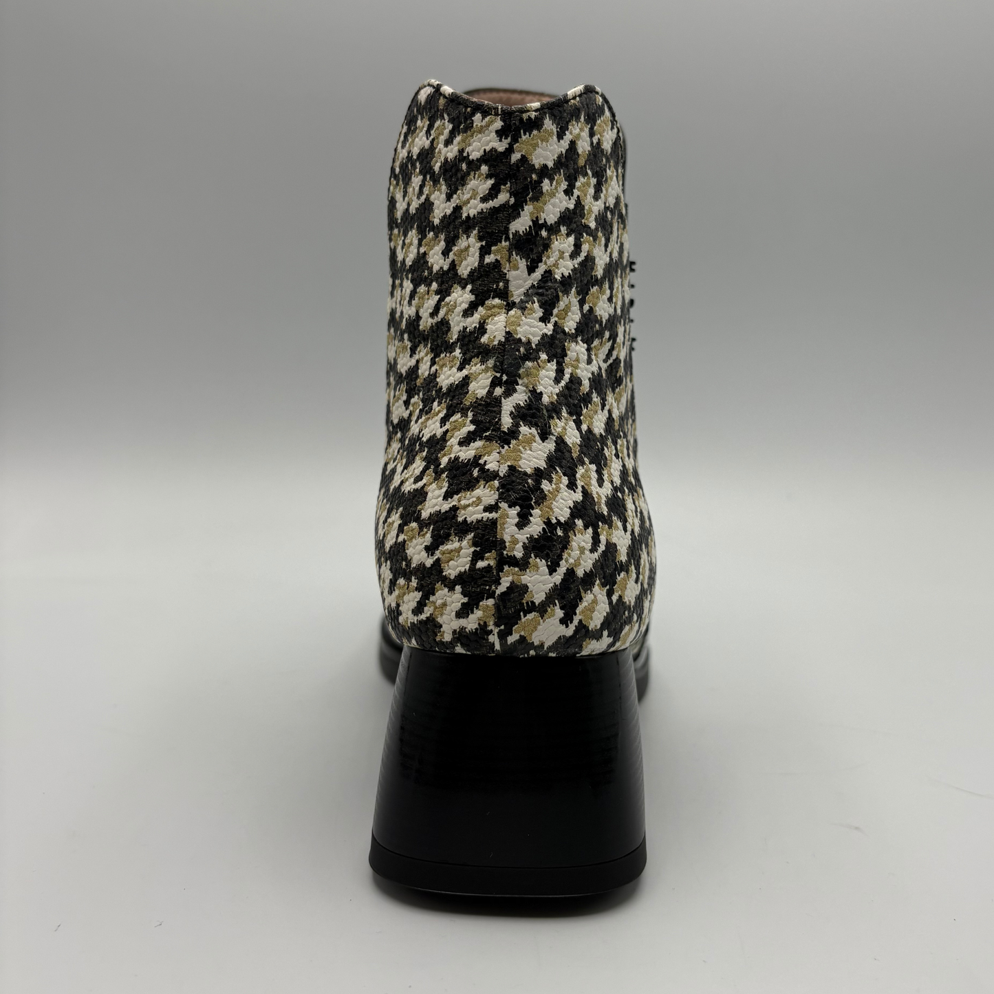 A single ankle boot shown from the back, featuring a black, beige, and white houndstooth patterned upper with a glossy black block heel, set against a neutral gray studio background.