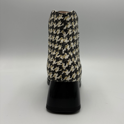 A single ankle boot shown from the back, featuring a black, beige, and white houndstooth patterned upper with a glossy black block heel, set against a neutral gray studio background.