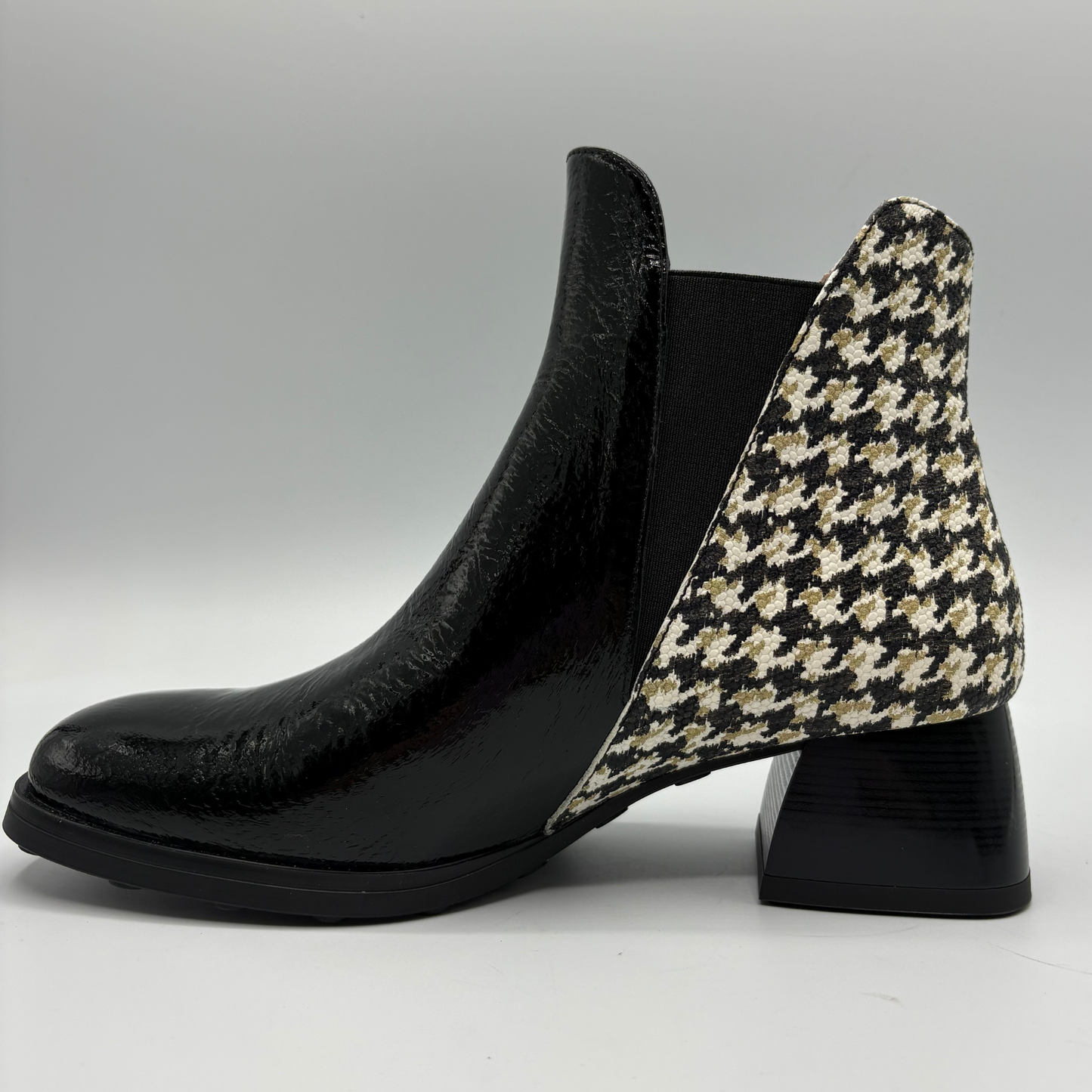 A black leather ankle boot with a black, beige, and white houndstooth patterned heel panel and a black elastic side gore, shown from a side profile on a light gray background.