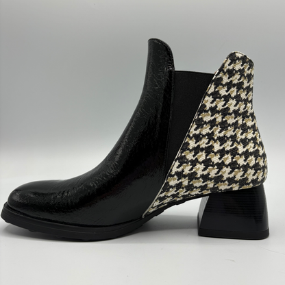 A black leather ankle boot with a black, beige, and white houndstooth patterned heel panel and a black elastic side gore, shown from a side profile on a light gray background.