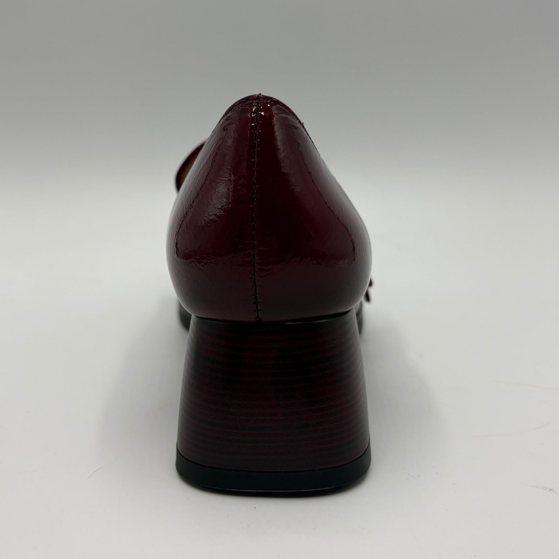 A single dark burgundy patent leather pump shown from the back, featuring a pointed toe, a block heel, and a black outsole, set against a plain light gray background.