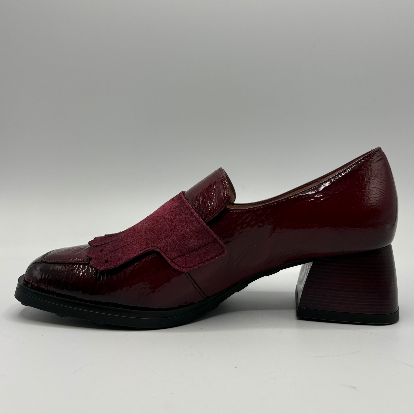 A single women's loafer-style shoe in deep burgundy, featuring a glossy patent leather toe cap, a suede-like side panel, and a chunky block heel, shown against a neutral gray background.
