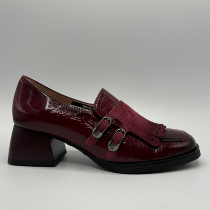 A single women's loafer-style shoe in deep burgundy, featuring a glossy patent leather upper, a suede tassel strap with silver-tone buckles, and a chunky block heel, photographed against a neutral gray background.