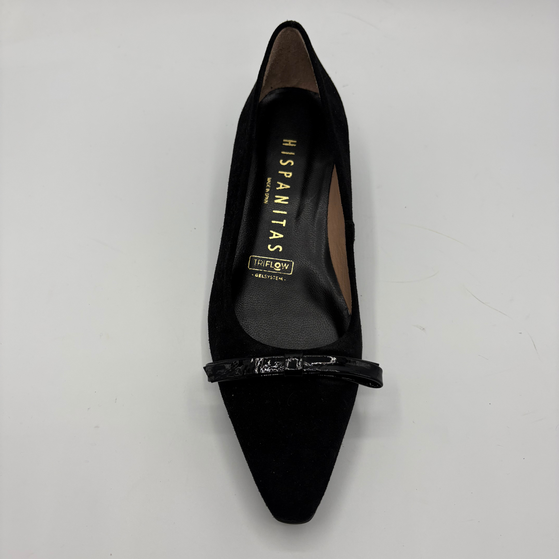 A single black pointed-toe flat shoe with a glossy black bow across the instep, photographed from a top-down angle on a light gray background. The insole displays the brand name 'HISPANITAS'.
