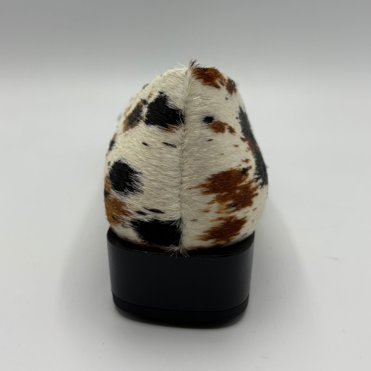 A single black-heel shoe shown from the back, featuring a cowhide upper with a white base and brown and black spots, set against a plain light gray background.