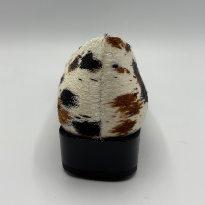 A single black-heel shoe shown from the back, featuring a cowhide upper with a white base and brown and black spots, set against a plain light gray background.