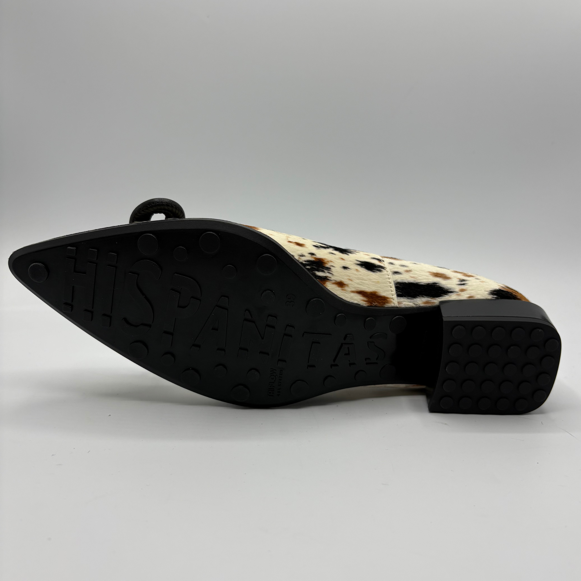 A single pointed-toe flat shoe shown from the side, featuring a black sole with 'HISPANITAS' embossed in large letters and a cream and brown cowhide-patterned upper.