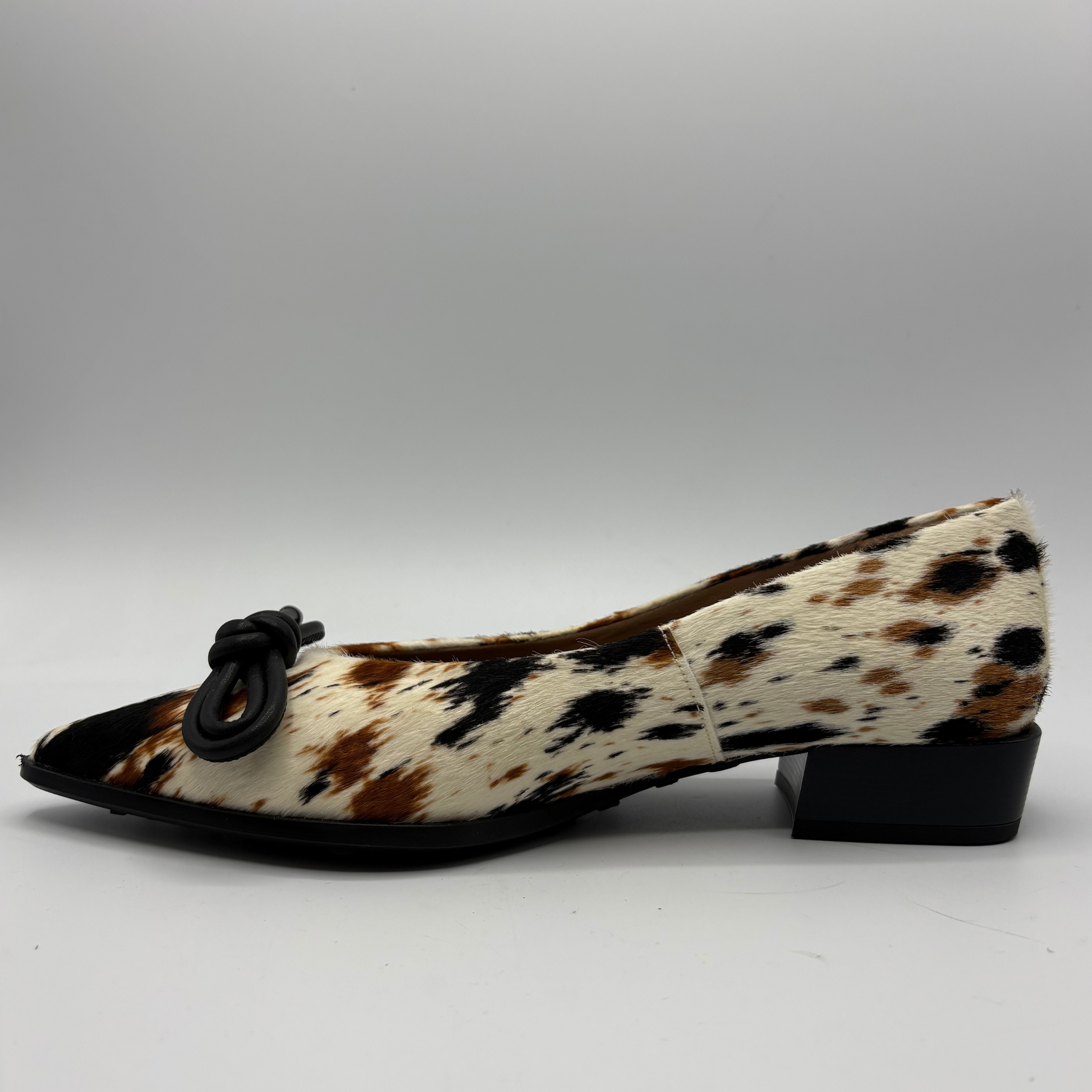 A single women's low-heel pump shoe featuring a brown, black, and cream cowhide pattern, finished with a black pointed toe and a black knotted bow detail on the vamp.