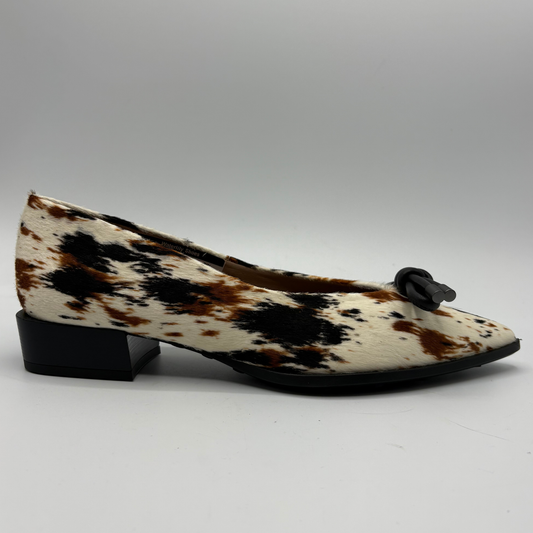 A single women's flat shoe with a black and brown cow print pattern on a cream base, featuring a low black block heel and a black bow detail on the toe, photographed on a neutral gray background.