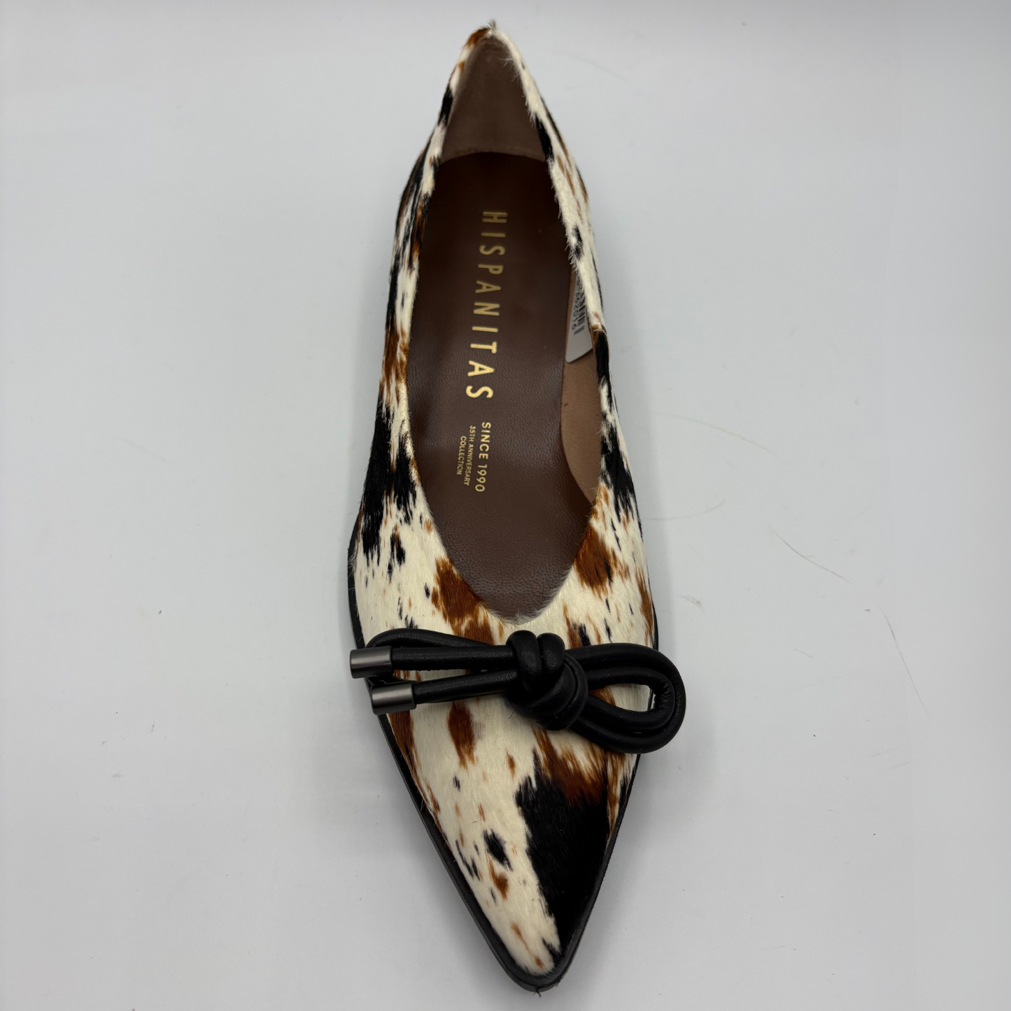 A pointed-toe flat shoe with a brown, black, and cream cowhide pattern, featuring a black fabric bow on the toe box. The insole displays the brand name 'HISPANITAS'.